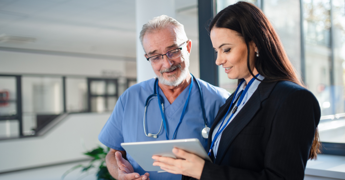 A doctor in scrubs and a woman in business attire look at a tablet together in a bright, modern building.